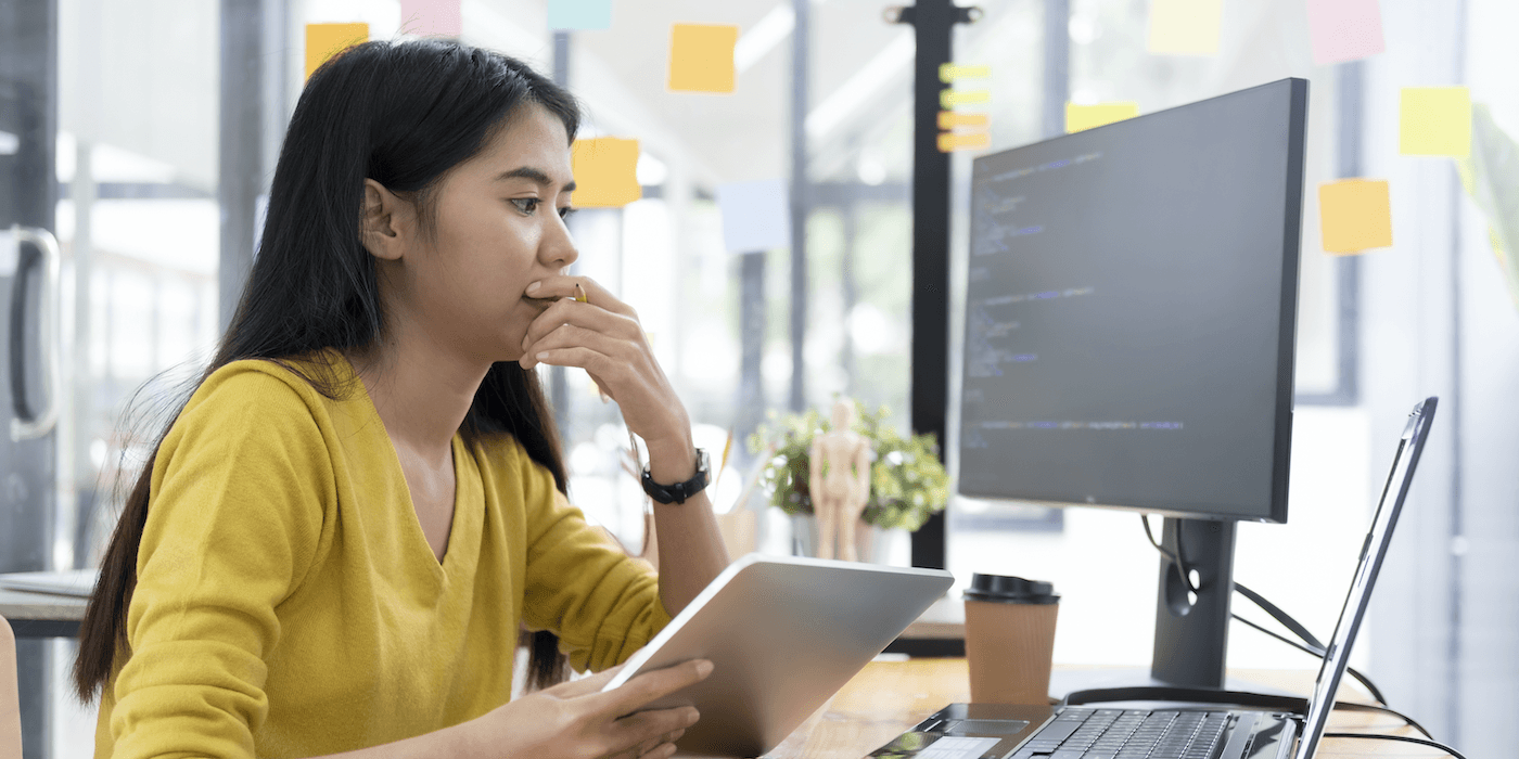 person sitting at desk looking at laptop and monitor screens showing software code