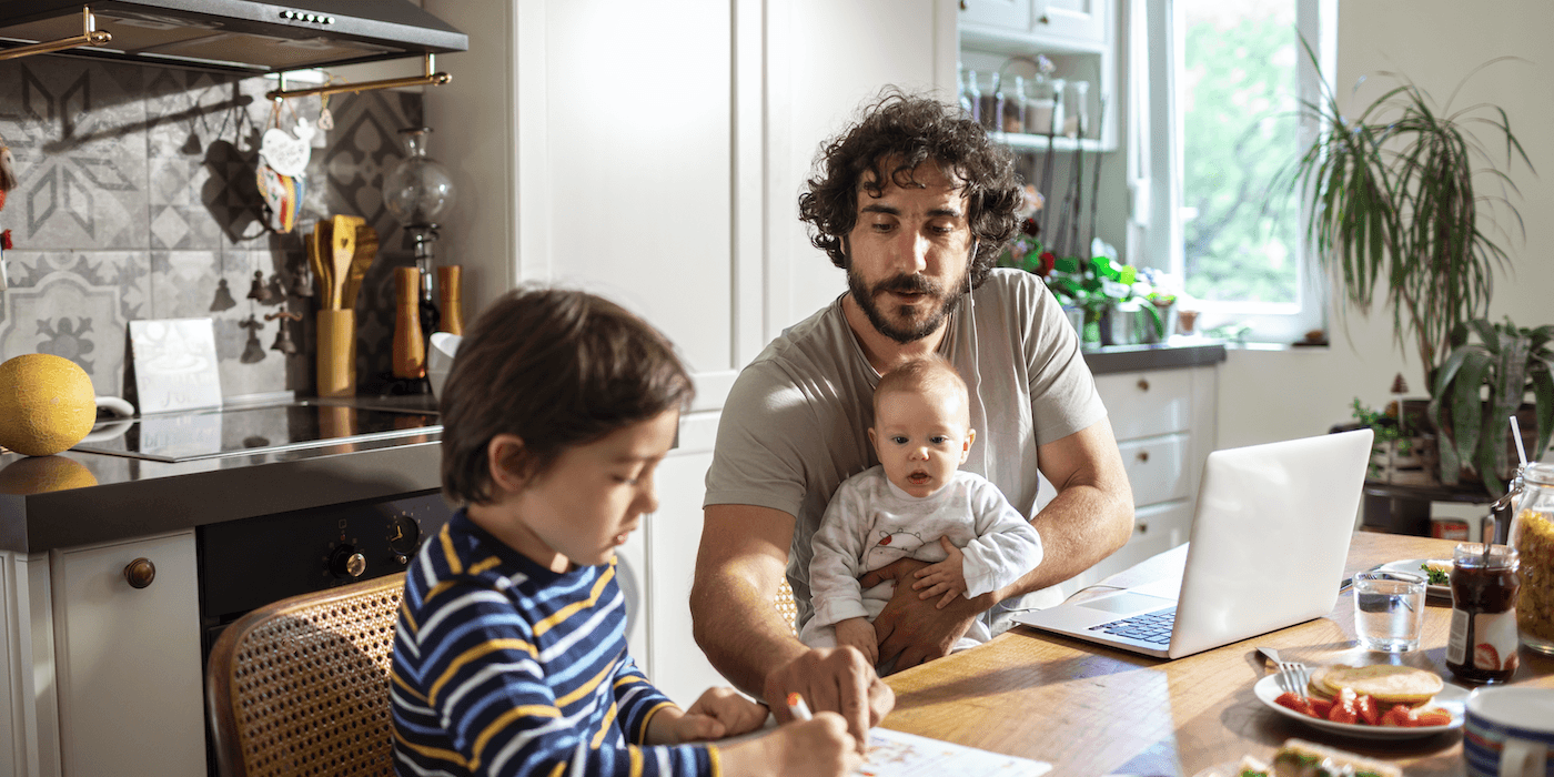 parent working at home with laptop open, a baby in their lap, and a child sitting next to them doing homework
