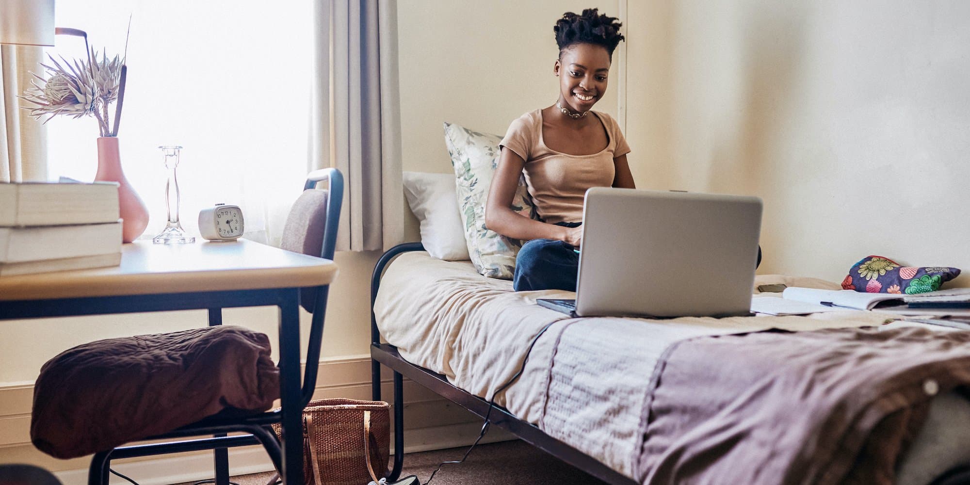 person sitting on bed in dorm room looking something up on a laptop and taking notes on a tablet
