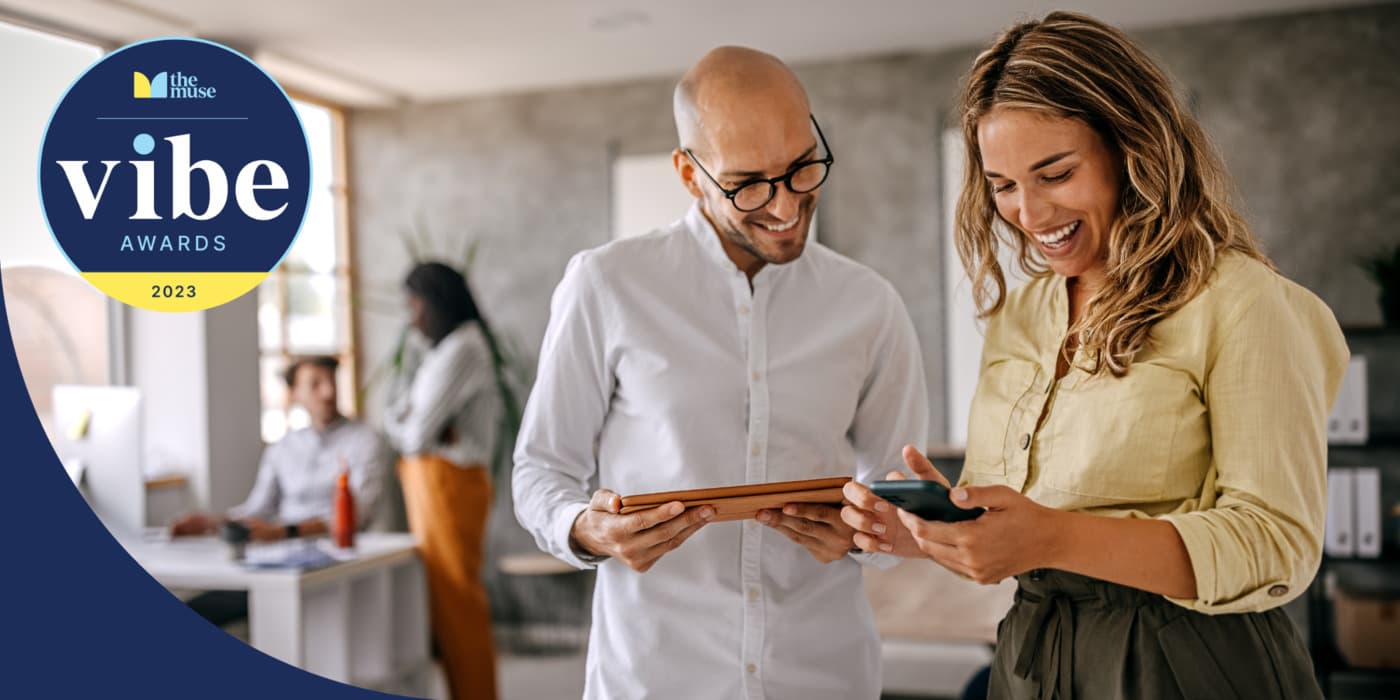 Two coworkers smiling while looking at a phone and tablet in an office.