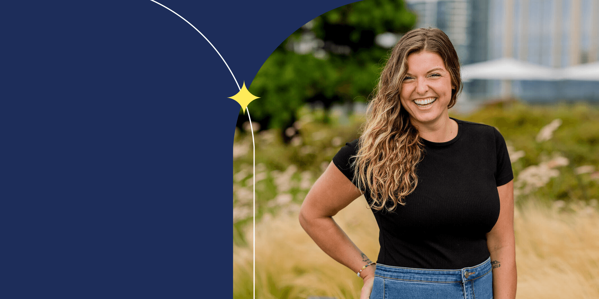 A smiling person facing the camera. They have long hair and wear a black shirt and blue jeans. An open field can be seen behind them.
