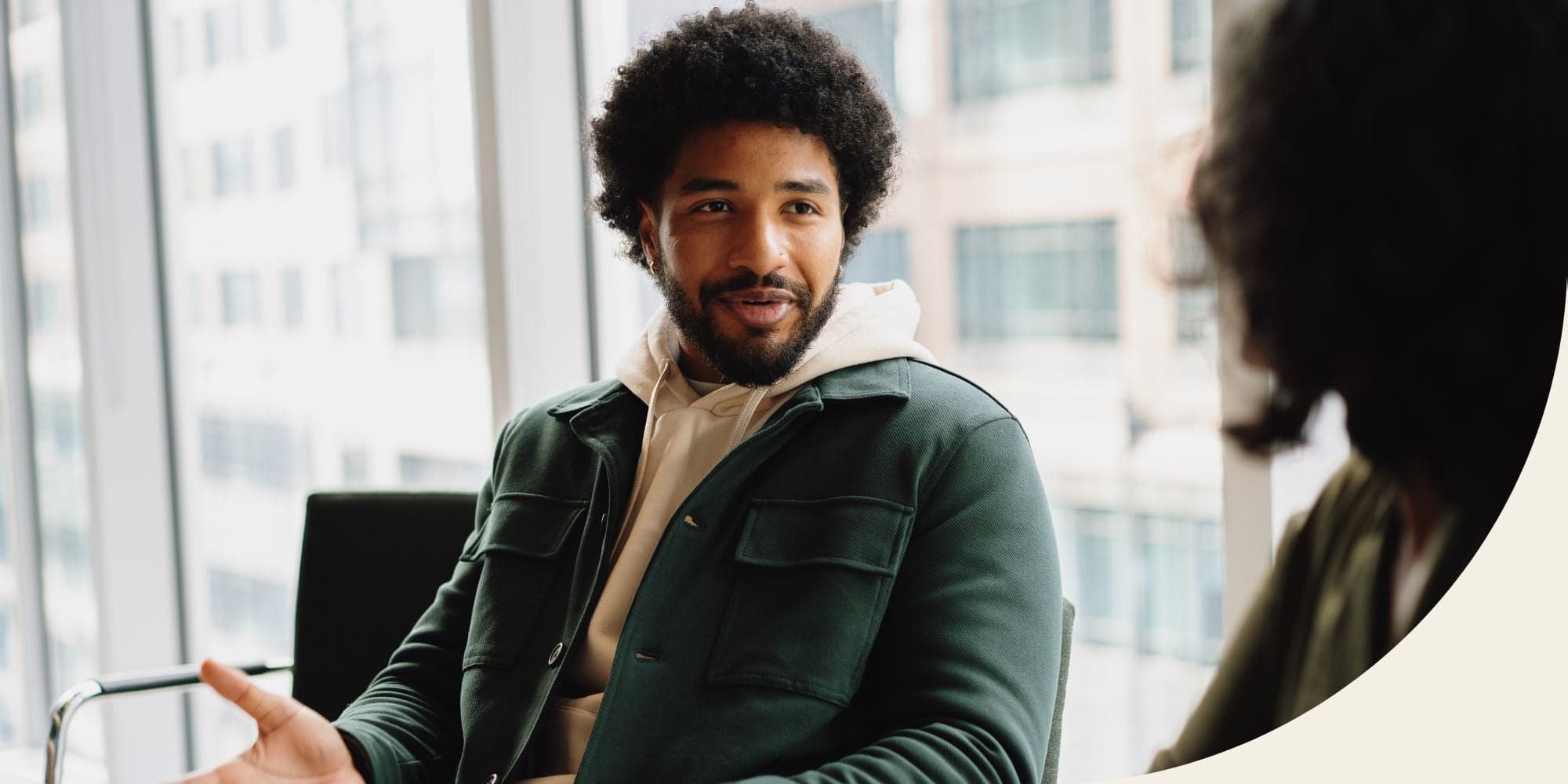 two people having a conversation in a conference room with large windows and buildings in the background; one person is facing the camera in a green jacket